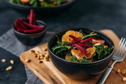 Wintersalat mit Grünkohl Mandarine und Rote Bete in schwarzer Schüssel auf Holzbrett neben Gabel und Pinienkernen vor dunklem Hintergrund.