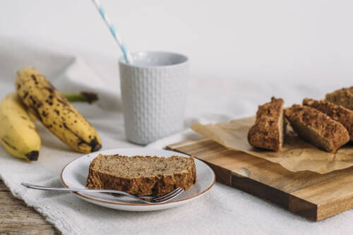 Zuckerfreies Bananenbrot auf Teller und Schneidebrett neben Bananen und Getränk vor hellem Hintergrund.